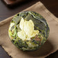 A round, tightly bound ball of loose-leaf tea, showing both dark green and pale yellow-green leaves, resting on a textured beige woven mat over a dark wooden surface.
