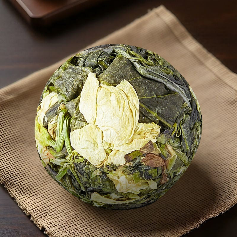 A round, tightly bound ball of loose-leaf tea, showing both dark green and pale yellow-green leaves, resting on a textured beige woven mat over a dark wooden surface.
