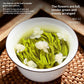 A white ceramic bowl filled with brewed green tea leaves and white flowers, placed on a woven mat. Text overlays describe the tea's color and appearance, with the brand name MO LI LONGZHU visible on the right.