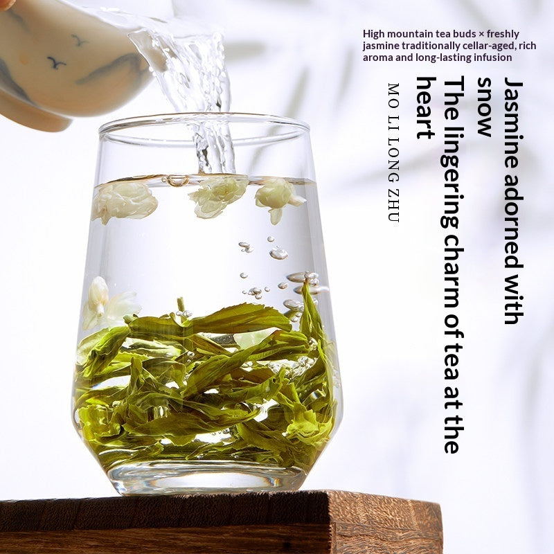 A clear glass tea infuser cup with green tea leaves and white flower buds inside, with hot water being poured into it from a ceramic pitcher. Text on the right describes jasmine-infused tea benefits, and the cup sits on a wooden surface against a white background.