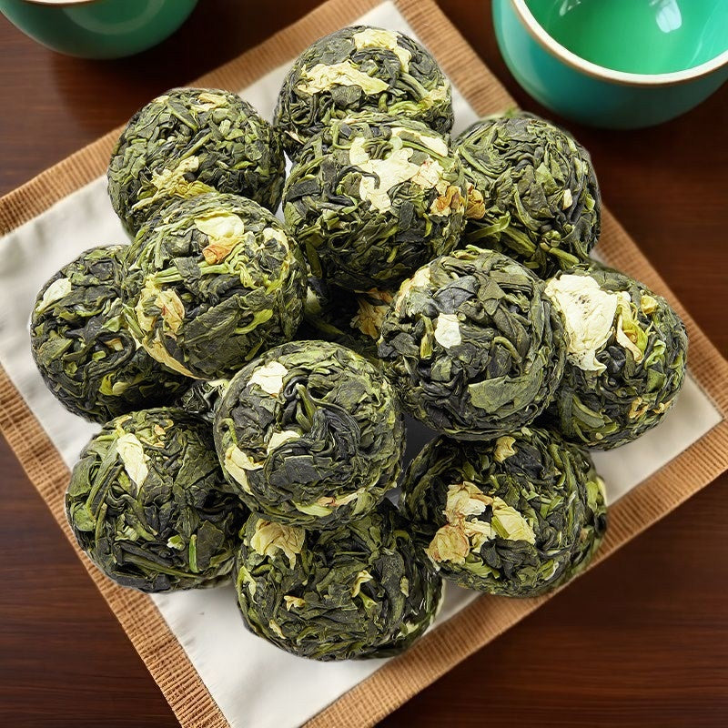 A pile of dark green tea cakes with visible white flower petals, arranged on a white paper liner inside a square bamboo tray on a dark wooden table, with teal cups partially visible in the background.