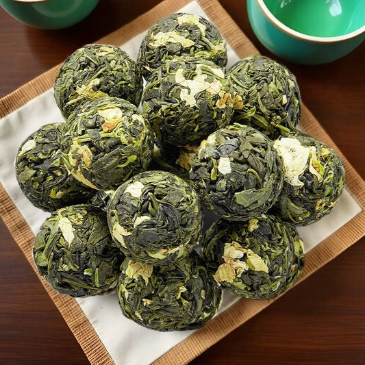 A pile of dark green tea cakes with visible white flower petals, arranged on a white paper liner inside a square bamboo tray on a dark wooden table, with teal cups partially visible in the background.