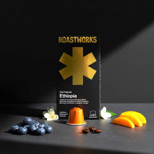Roastworks coffee packaging with a coffee pod and fruits on a dark surface